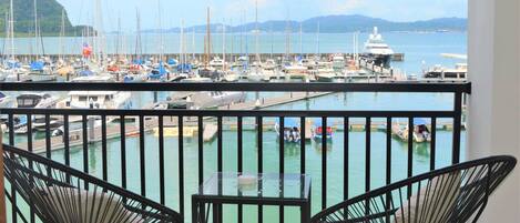 Un balcón con dos sillas negras y una mesita pequeña, con vista a un puerto deportivo con varias embarcaciones y una montaña distante.