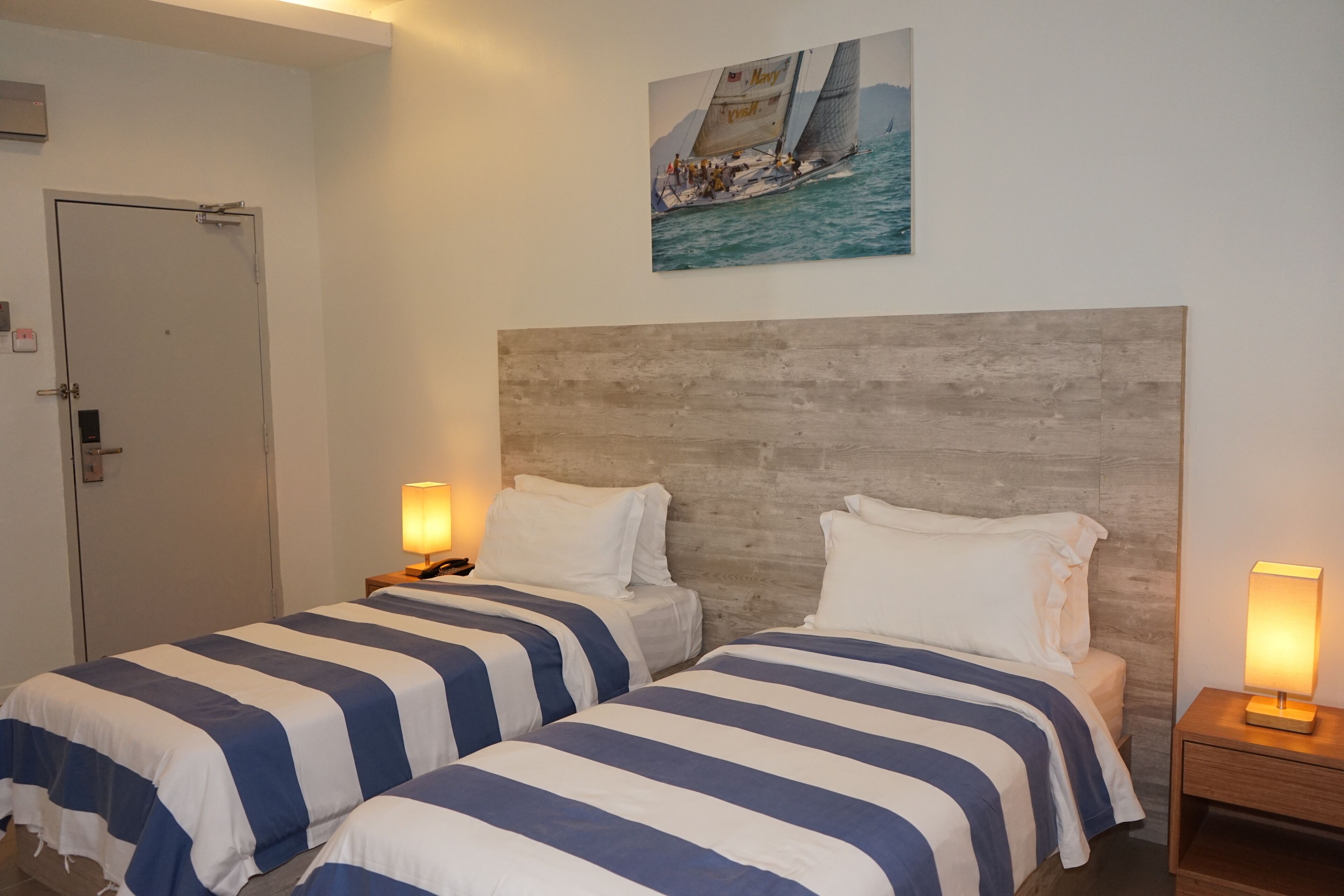 Une chambre d’hôtel avec deux lits, une tête de lit en bois, un tableau représentant un voilier et une porte.