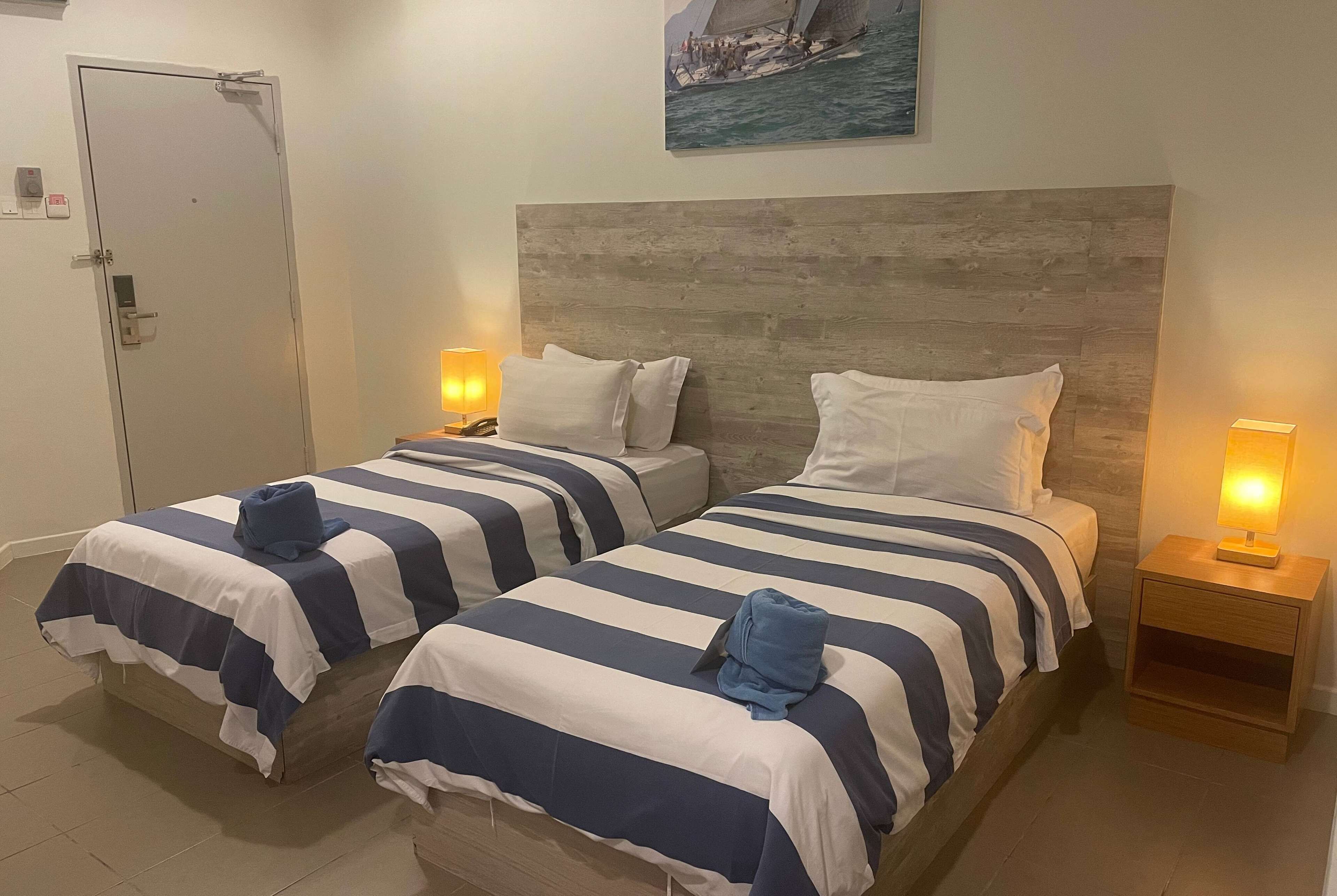 Una habitación de hotel con dos camas, cada una con colcha y almohadas a rayas, un cabecero de madera, lámparas de noche y un cuadro de un velero en la pared.