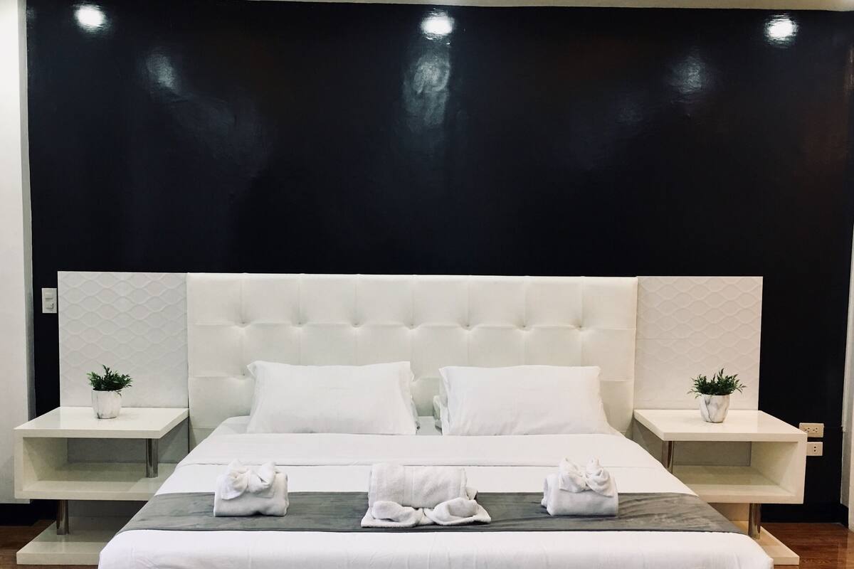 Une chambre d’hôtel moderne avec un grand lit, une tête de lit blanche et deux tables de chevet ornées de plantes en pot.