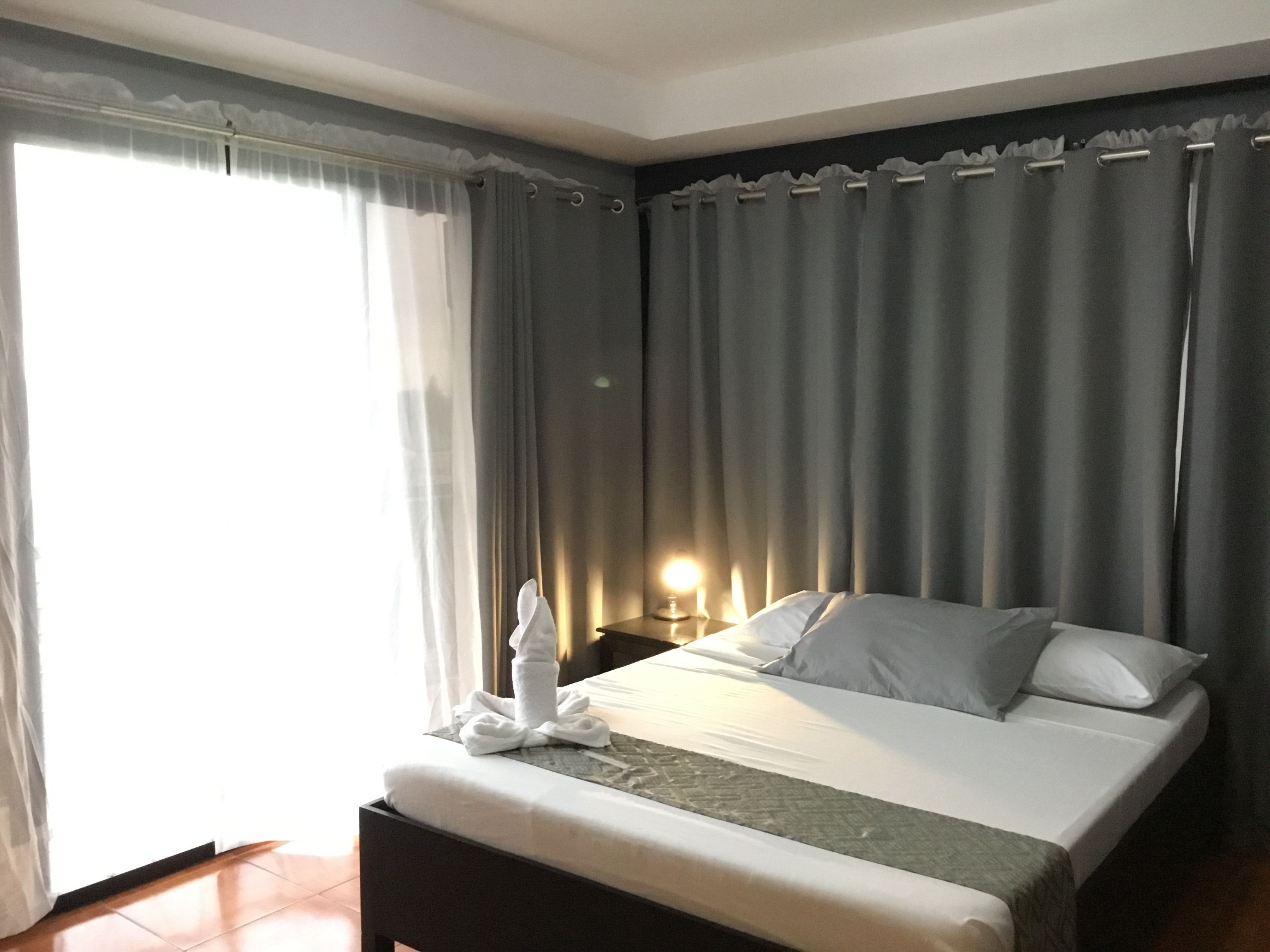 A hotel room with a large bed, a bedside table with a lamp, and a view of the outside through sheer curtains.