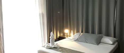 A hotel room with a large bed, a bedside table with a lamp, and a view of the outside through sheer curtains.