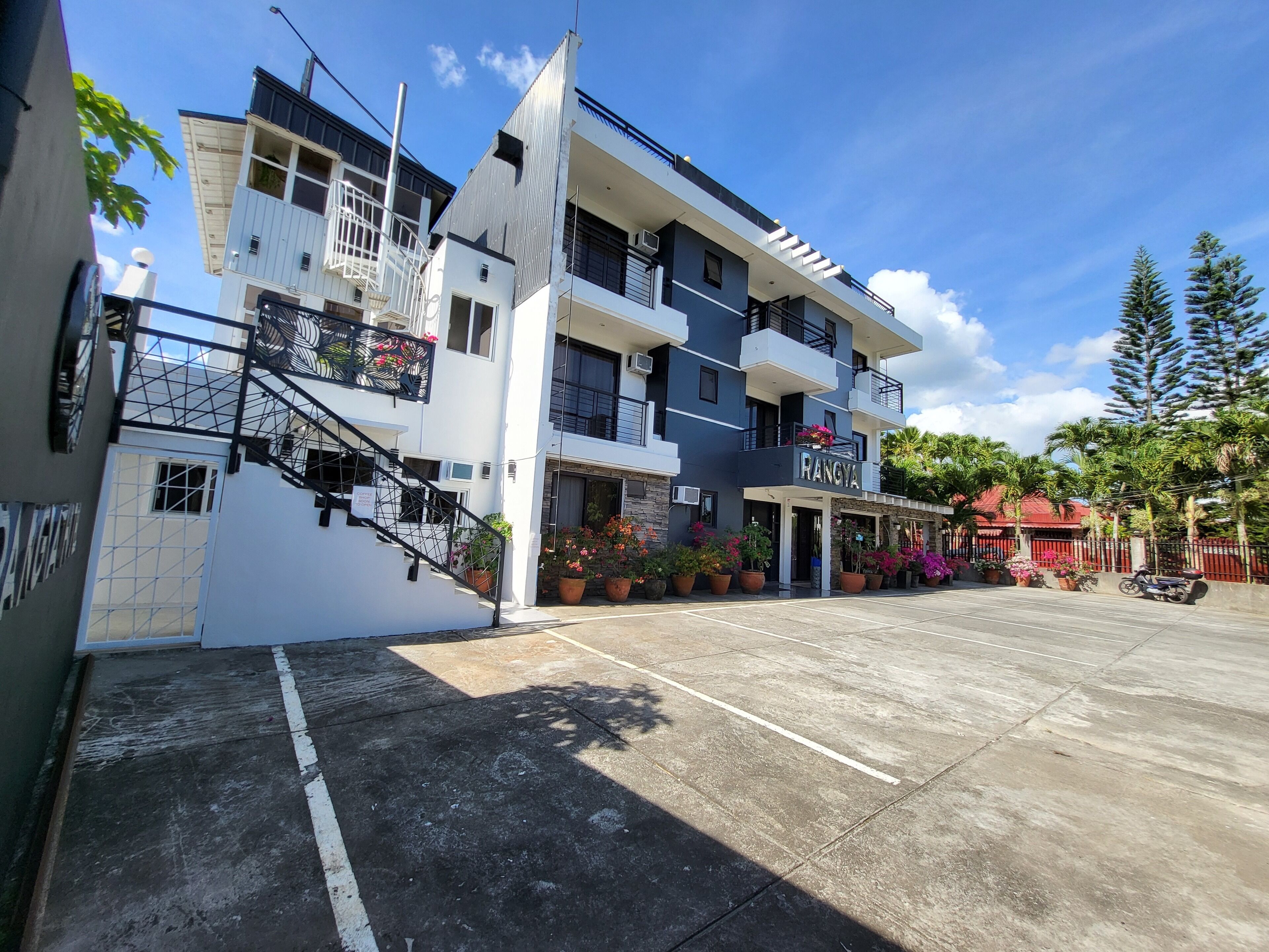 A multi-story building with a modern design, featuring balconies and a parking area in front.