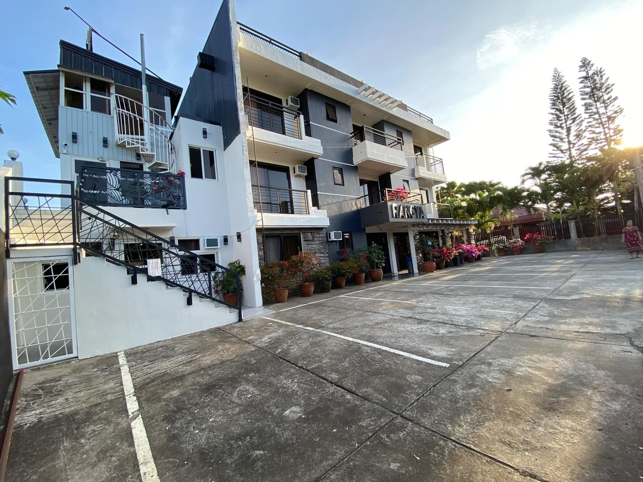A multi-story hotel with a modern design, featuring a prominent staircase, balconies, and a parking area.