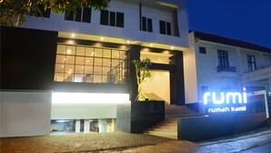A modern building with a large illuminated entrance, a staircase leading up to the main entrance, and a sign that reads "rumi rumah kami".