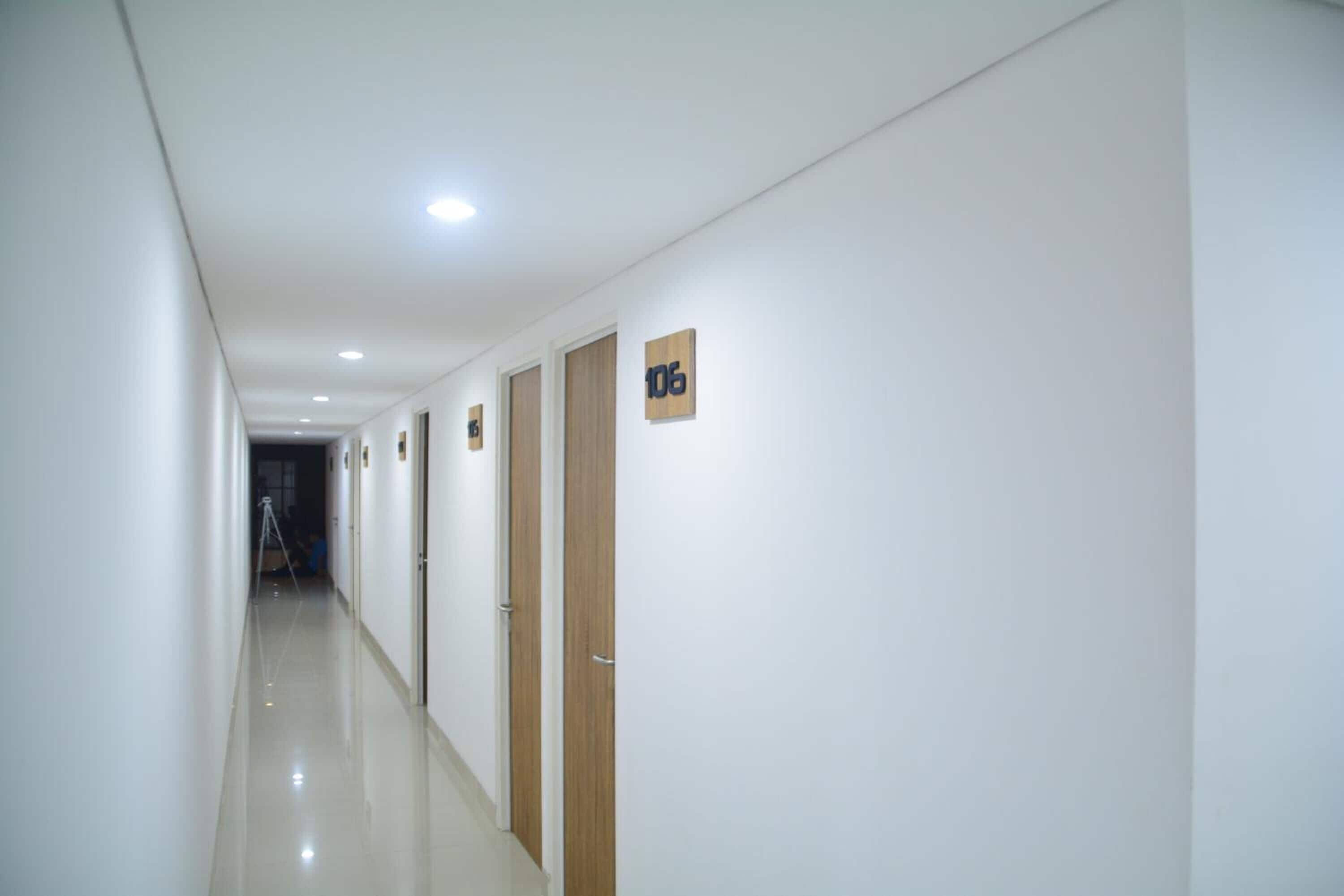 Um corredor comprido e estreito com paredes brancas e piso brilhante. Há portas no lado direito, cada uma delas com um número.
