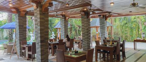 A restaurant with wooden pillars, tables, and chairs, and a view of greenery outside.