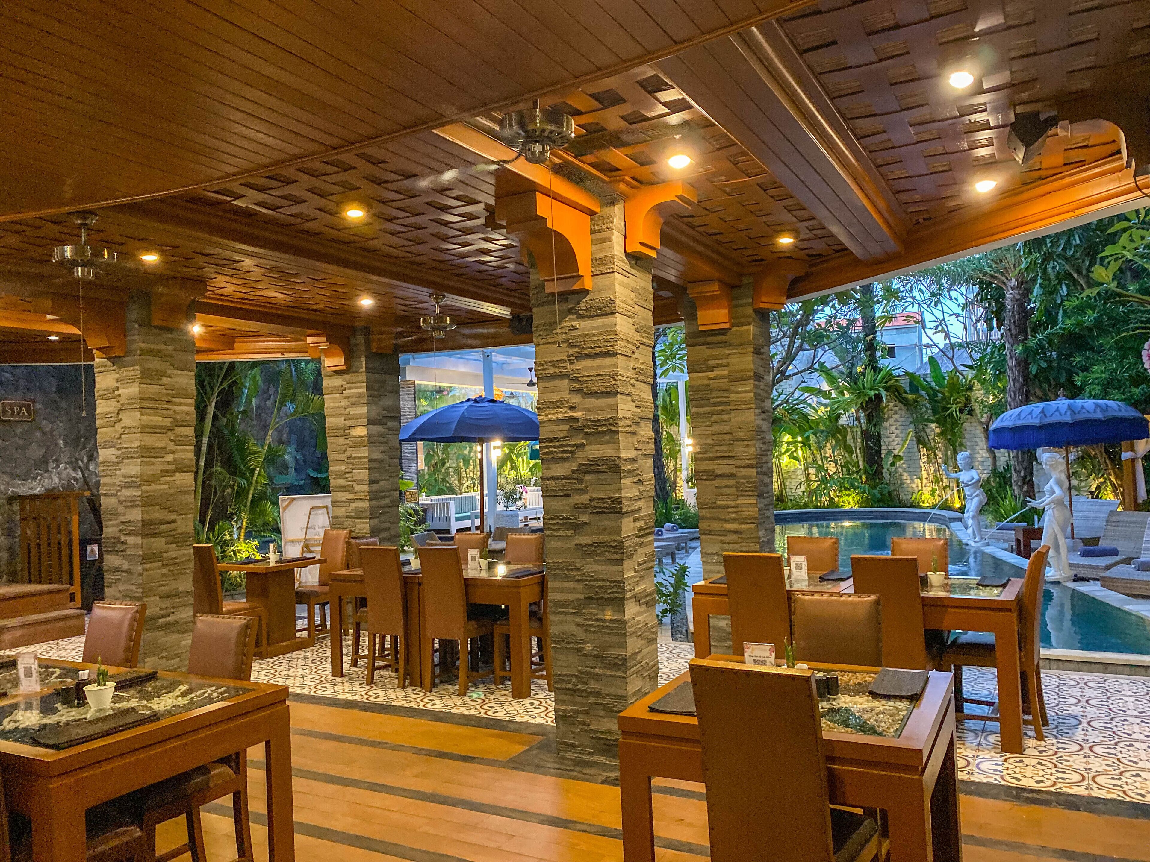 A restaurant with stone pillars, wooden tables, and chairs, and a pool area with umbrellas.