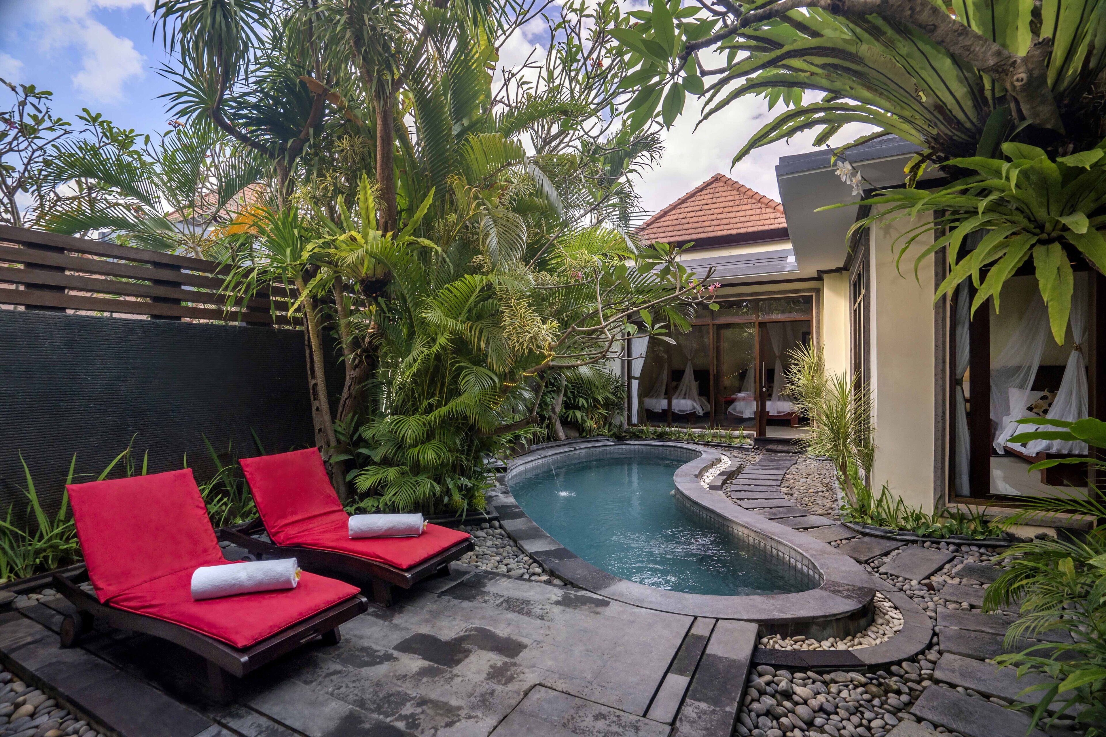 Un área de piscina privada con dos sillones rojos, rodeada de plantas tropicales y un sendero de piedra que lleva a una casa con techo de tejas.