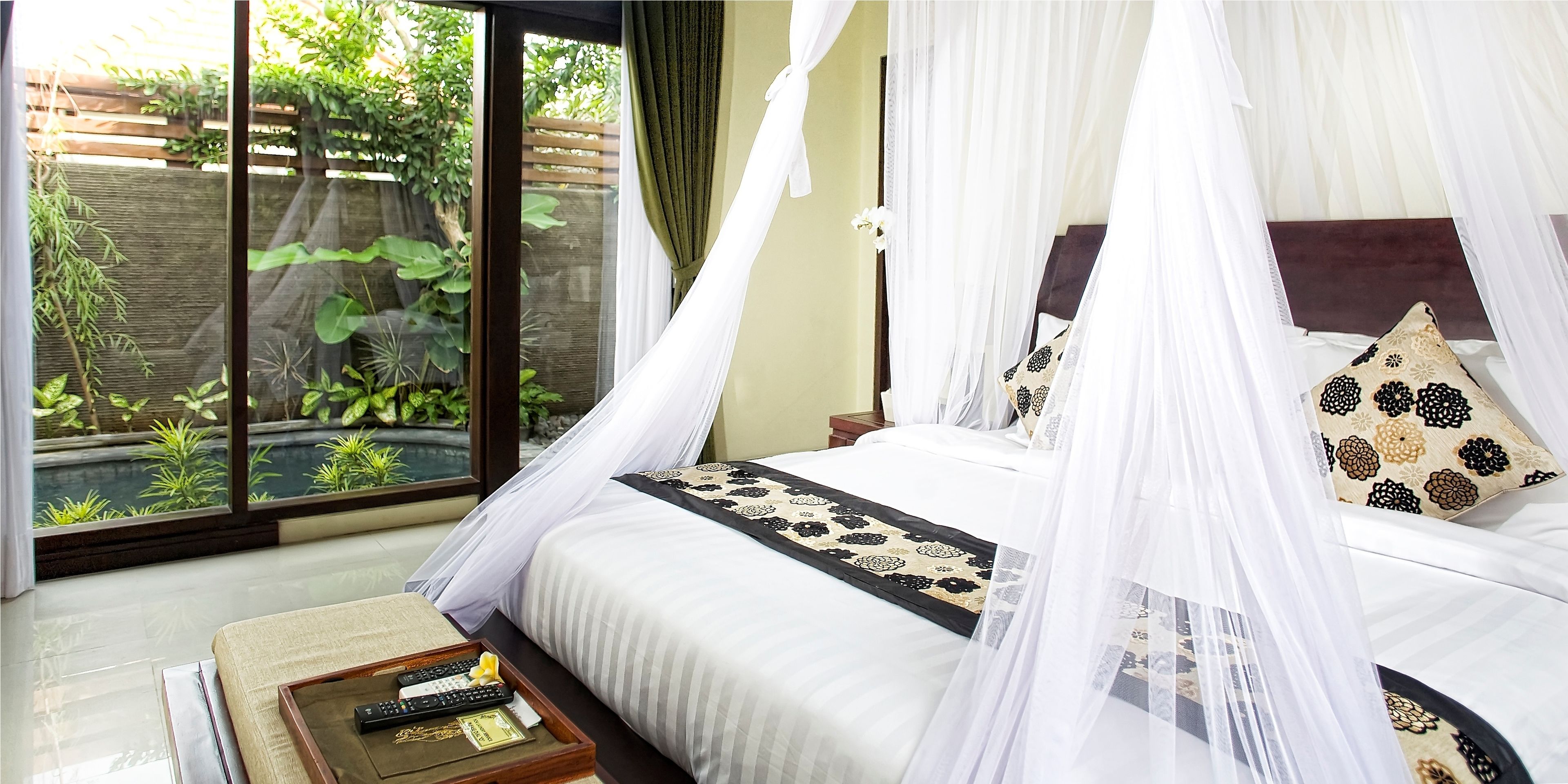 A bedroom with a bed under a canopy, a bedside table with a tray, and a view of greenery through a window.