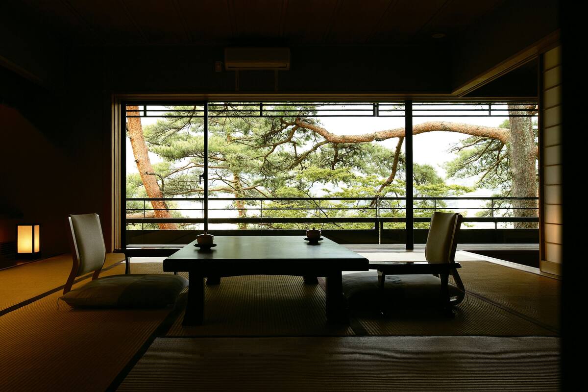 Una habitación japonesa tradicional con suelo de tatami, una mesa baja y sillas. Un ventanal muestra un árbol fuera.