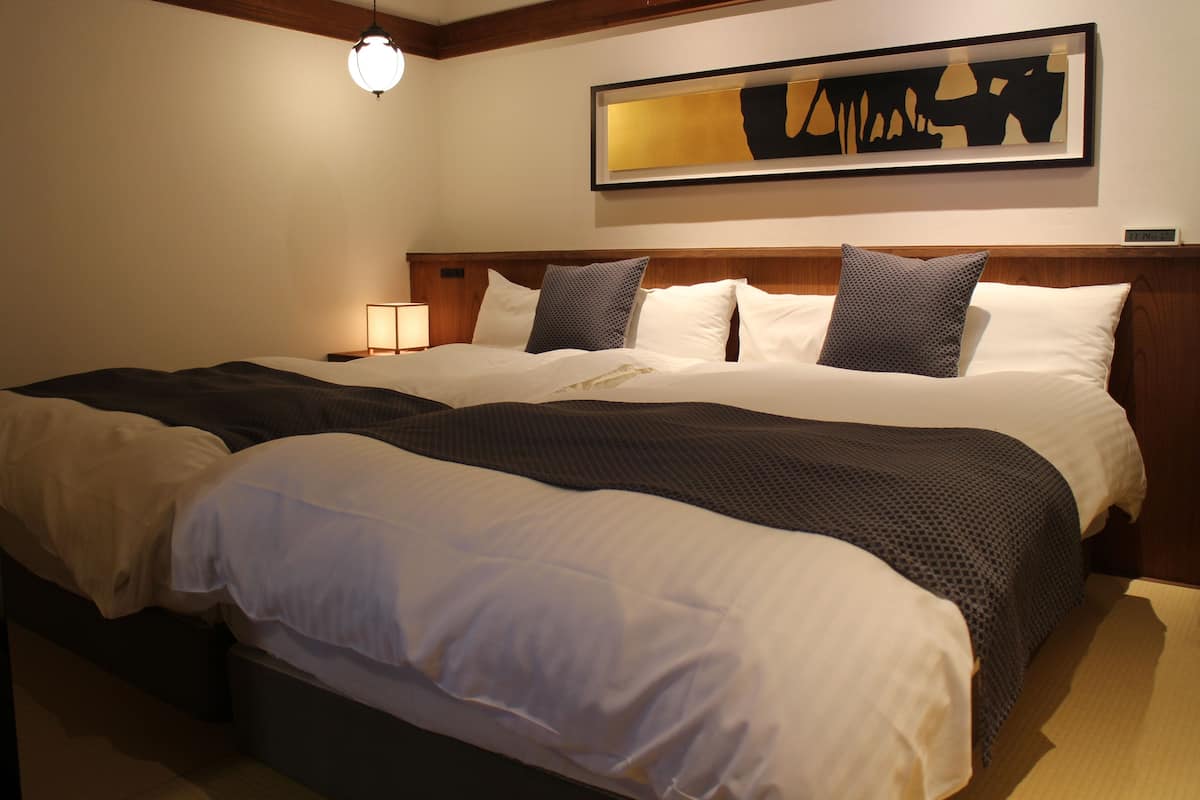 Una cama doble con cabecera de madera, ropa de cama blanca y con estampado oscuro, y una mesita de noche con una lámpara.