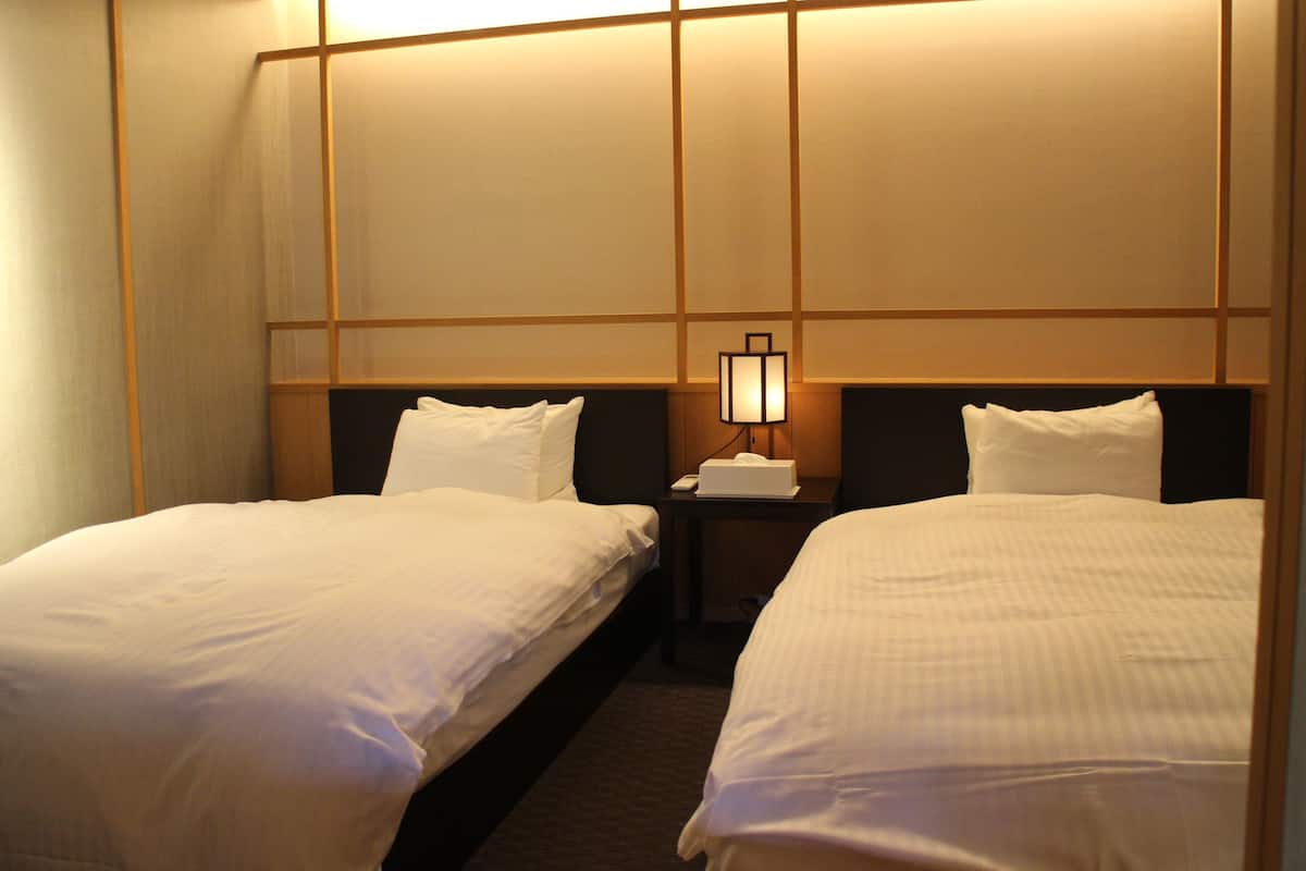 Habitación de hotel con dos camas individuales, una mesita de noche con lámpara y un ventanal con vistas al horizonte de la ciudad.