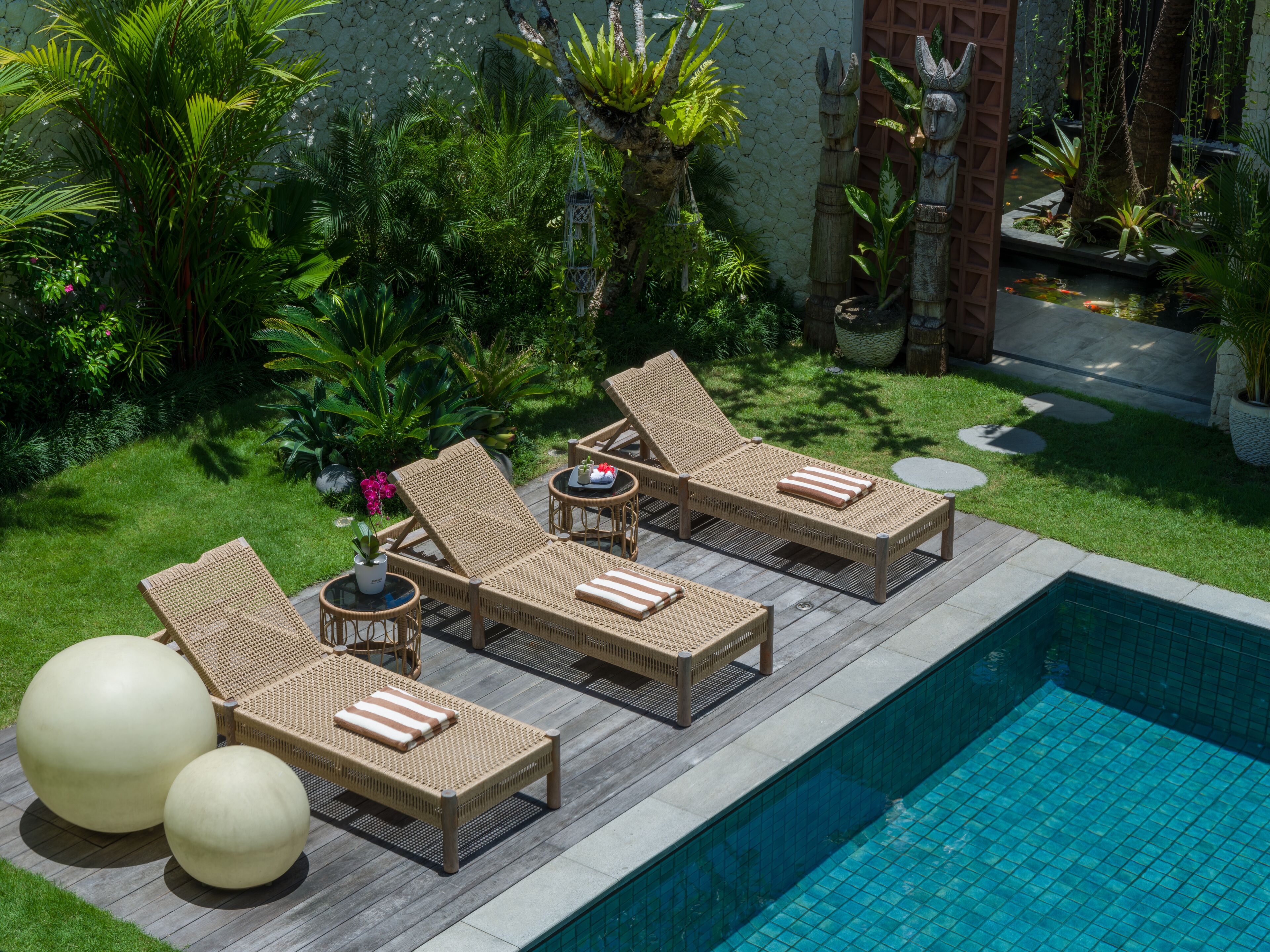 Zona de piscina con sillones reclinables y una pequeña mesa, rodeada de exuberante vegetación y un muro de piedra.