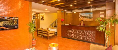 A hotel lobby with a reception desk, a TV mounted on the wall, a sofa, a coffee table, and potted plants.