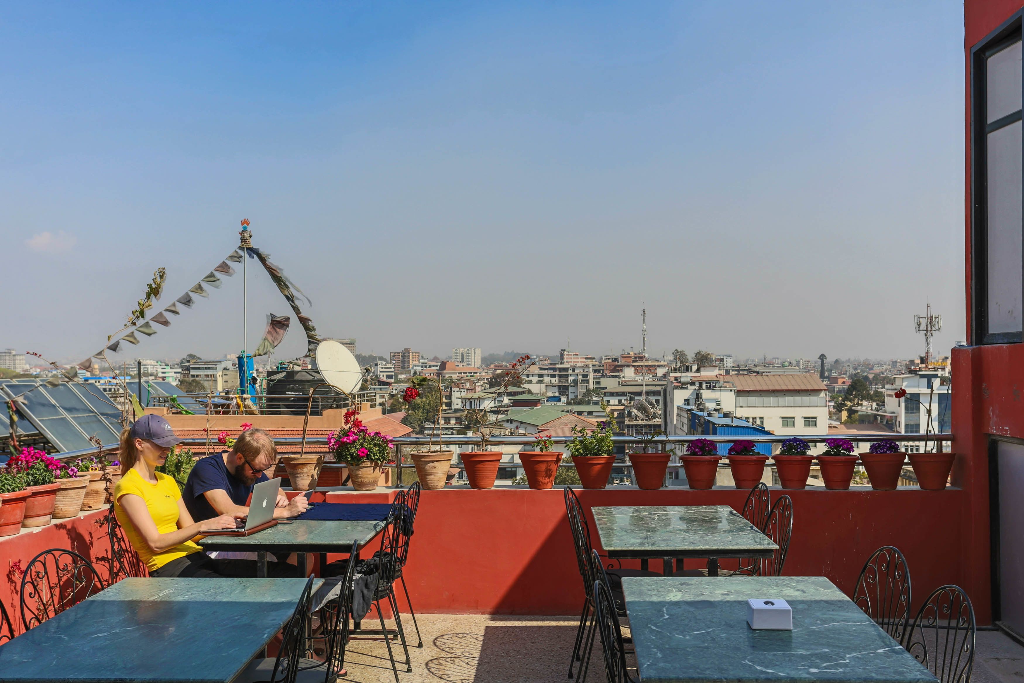 A rooftop terrace with tables and chairs, potted plants, and a view of the cityscape.