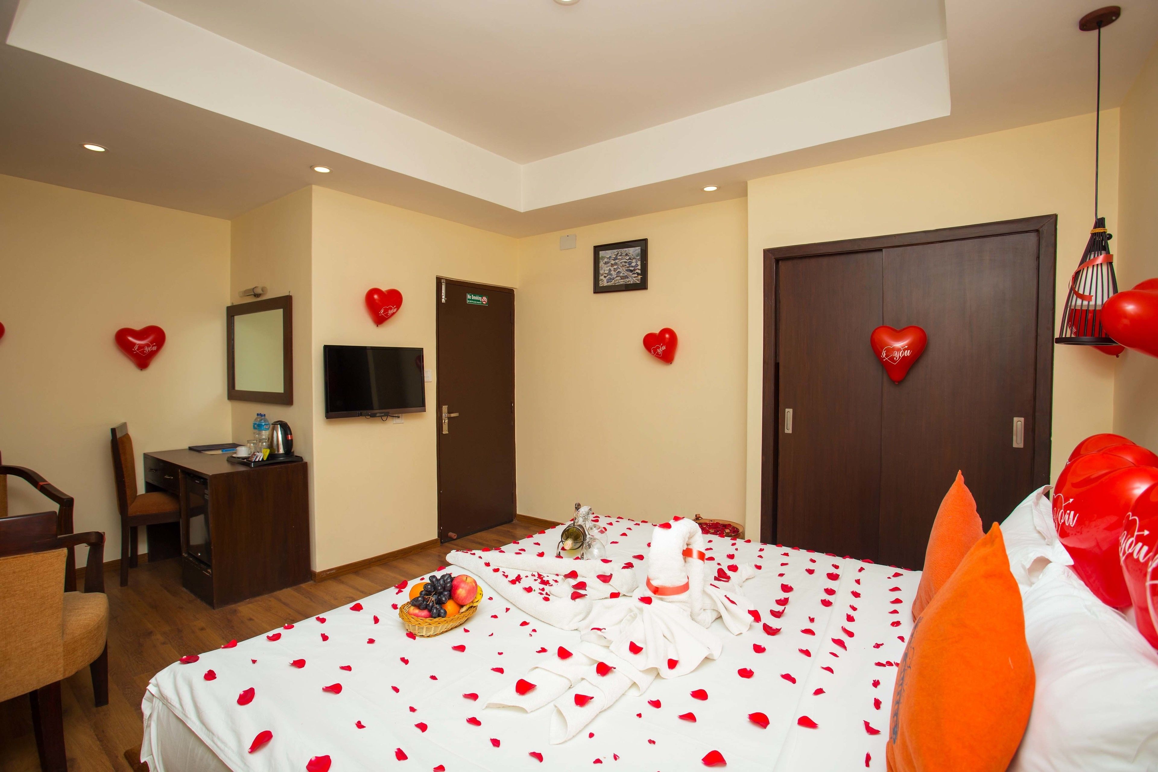 Una camera d'albergo con un letto ricoperto di petali di rosa, una scrivania con un computer, una TV e decorazioni a forma di cuore sulle pareti.