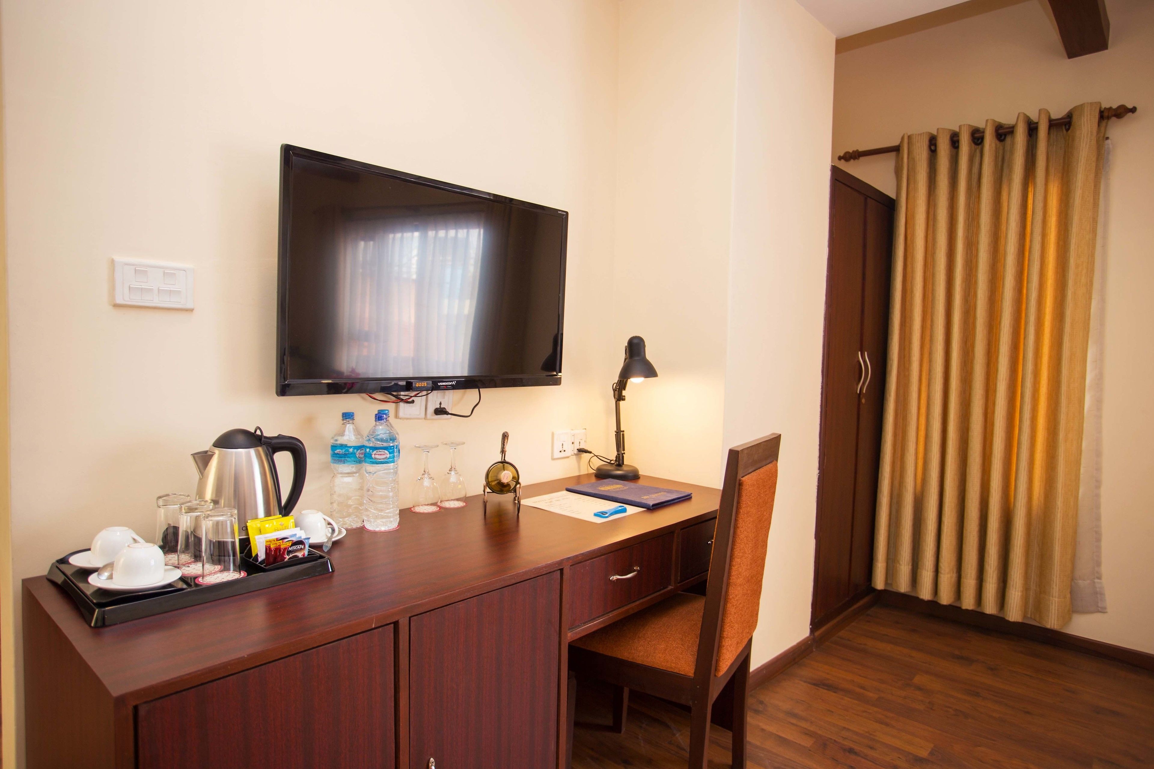 A hotel room with a flat-screen TV mounted on the wall, a desk with a lamp, a chair, and a wooden floor.