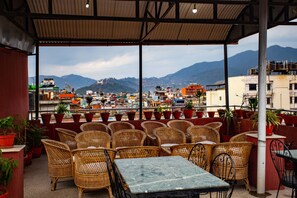 A covered outdoor seating area with wicker chairs and tables, overlooking a cityscape with mountains in the background. - Hotel Jay Suites (Kathmandu)