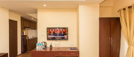 Quarto de hotel com cozinha compacta, TV fixada na parede, guarda-roupa de madeira e uma pequena escrivaninha com um cesto de lixo.