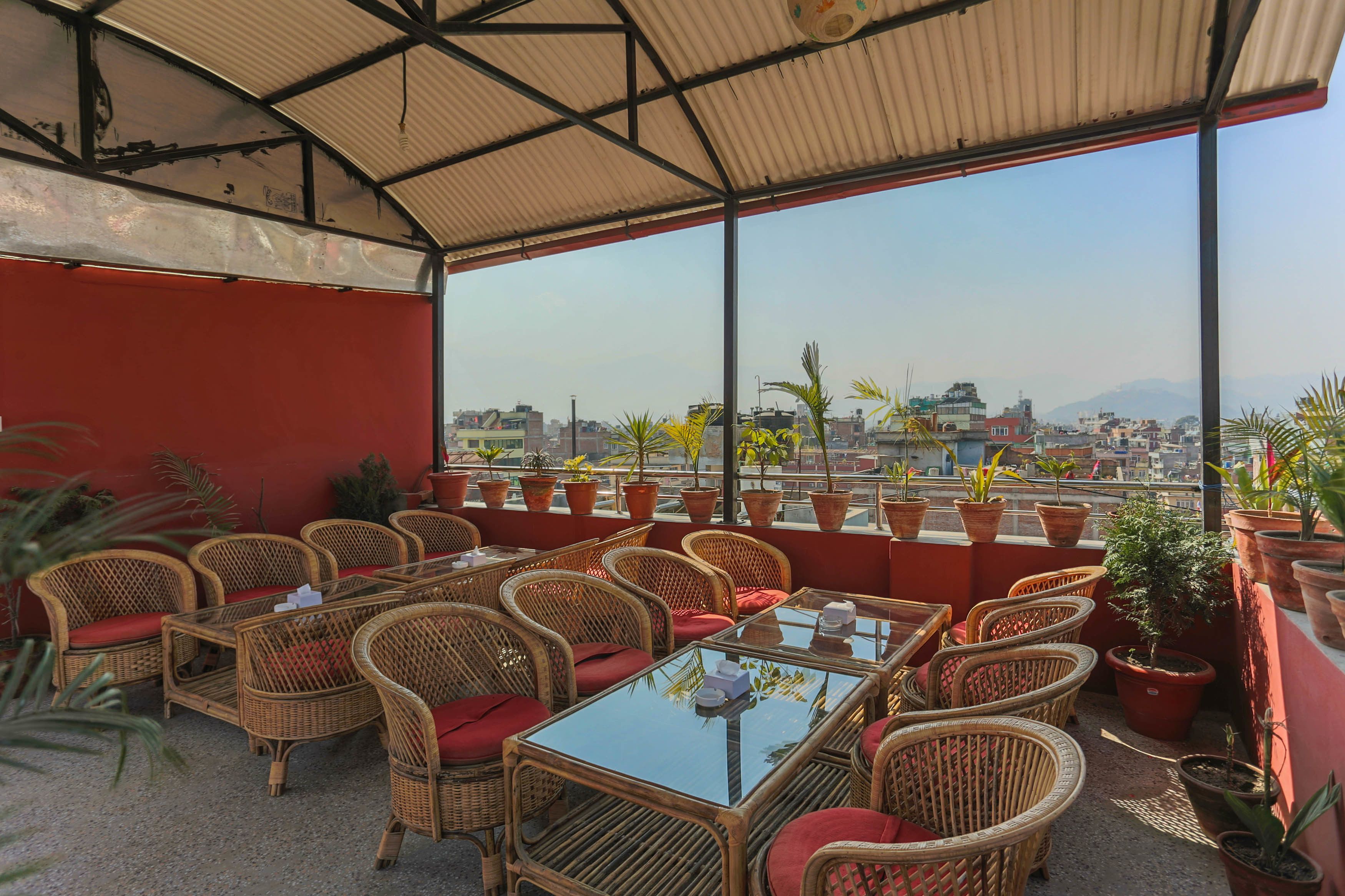 An outdoor seating area with wicker furniture and red cushions, overlooking a cityscape with buildings and mountains.
