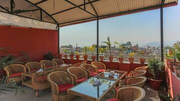 Un espacio al aire libre con mobiliario de mimbre y cojines rojos, con vistas a un paisaje urbano que incluye edificios y montañas.