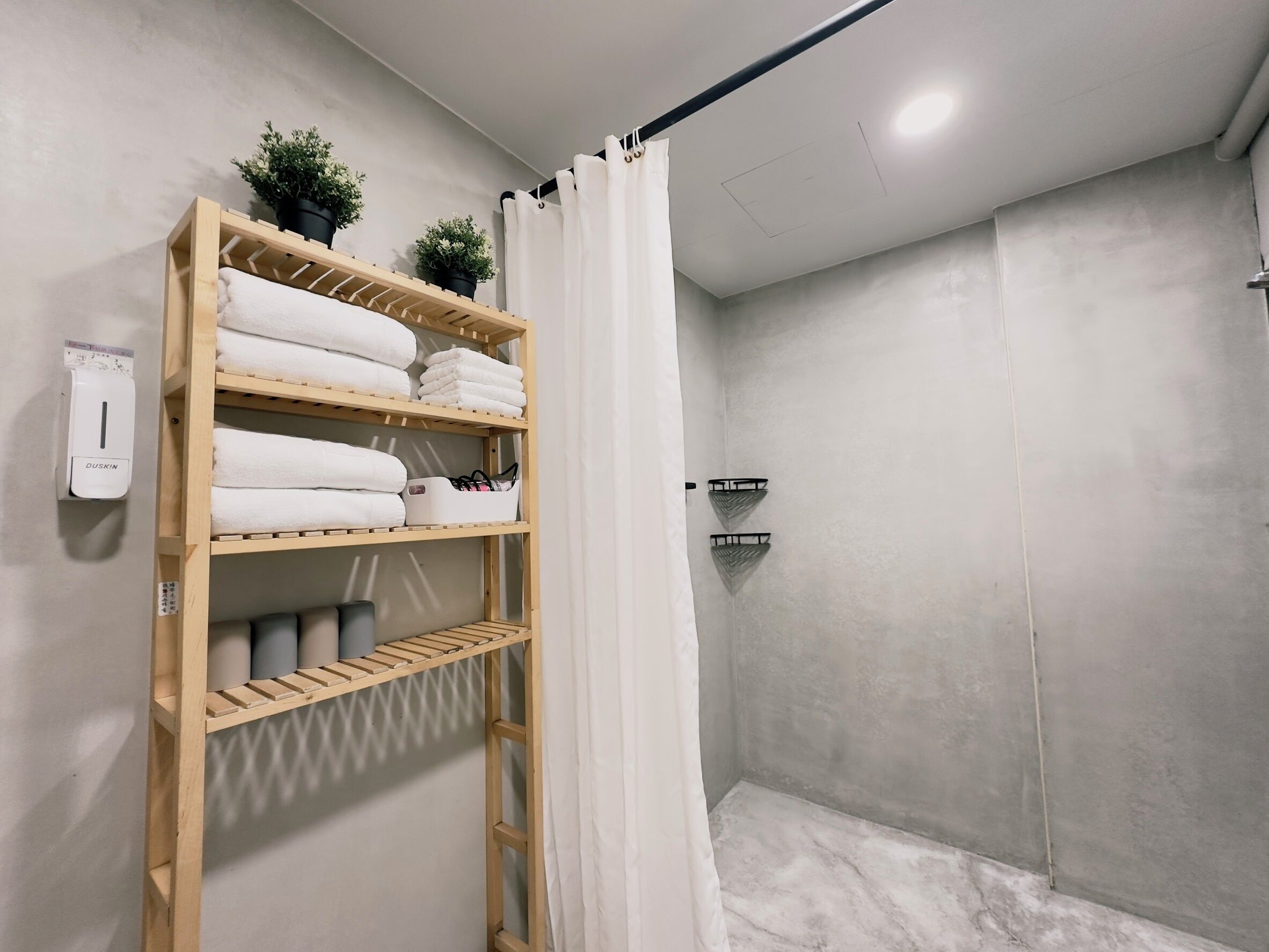 Un baño con estante de madera que contiene toallas, una cortina de ducha y un dispensador de jabón en la pared.