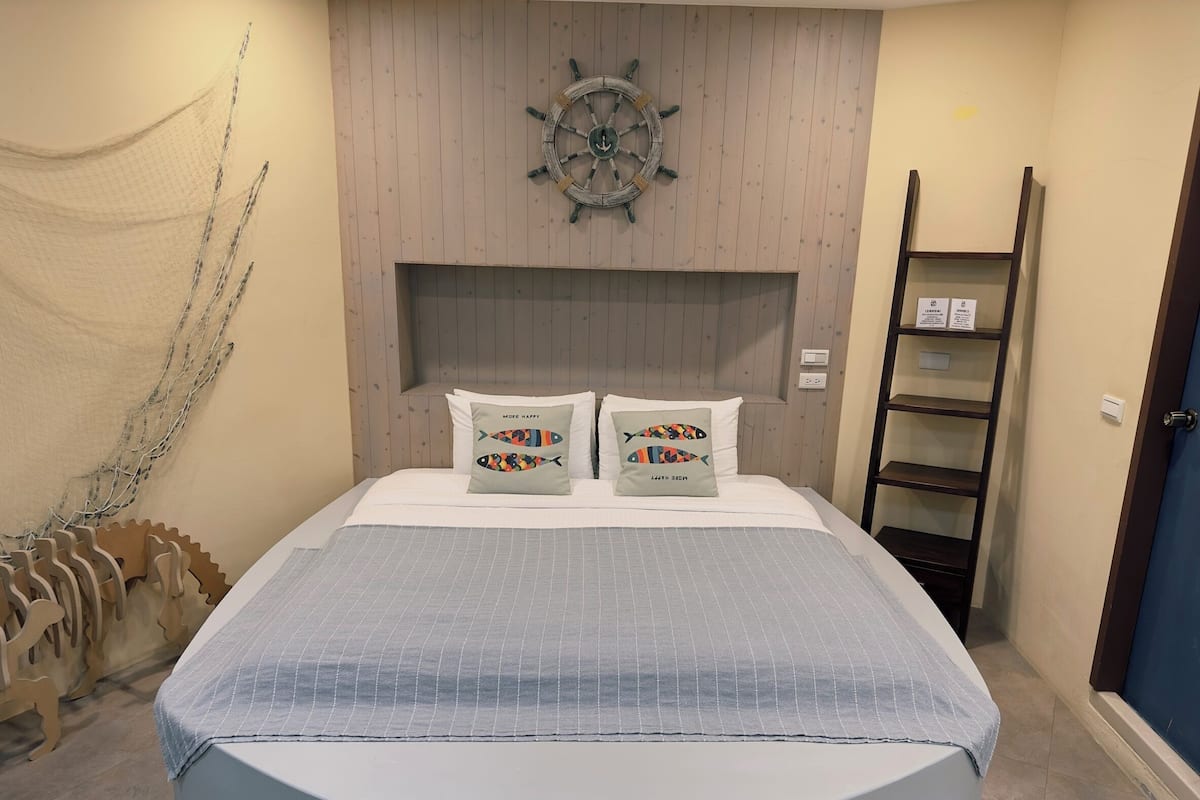 A bedroom with a bed, two pillows, a headboard, a wall-mounted ship's wheel, a wooden ladder, and a shelf with items.