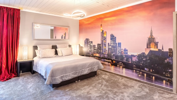 A bedroom with a cityscape mural, a bed with white linens, a nightstand, and a red curtain. - PMC Business Apartments (Ruesselsheim)