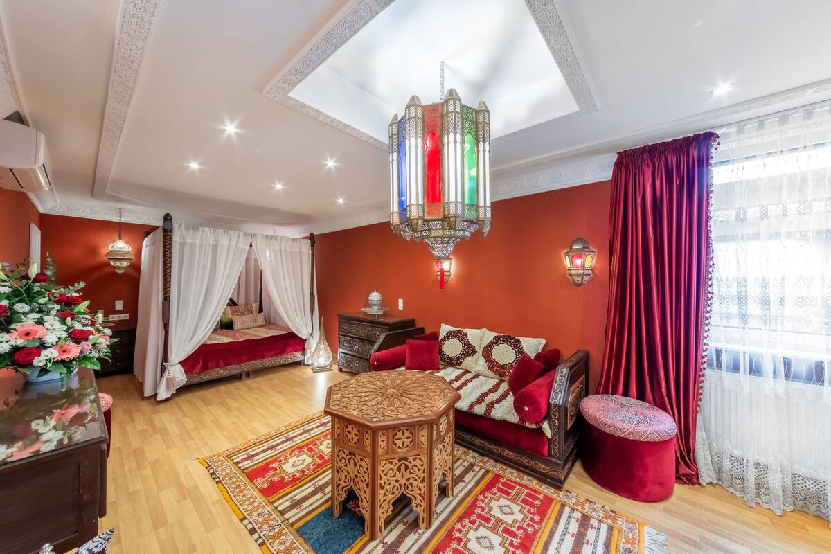 Quarto com cama, mesa de cabeceira de madeira, banquinho vermelho, tapete estampado e lustre.