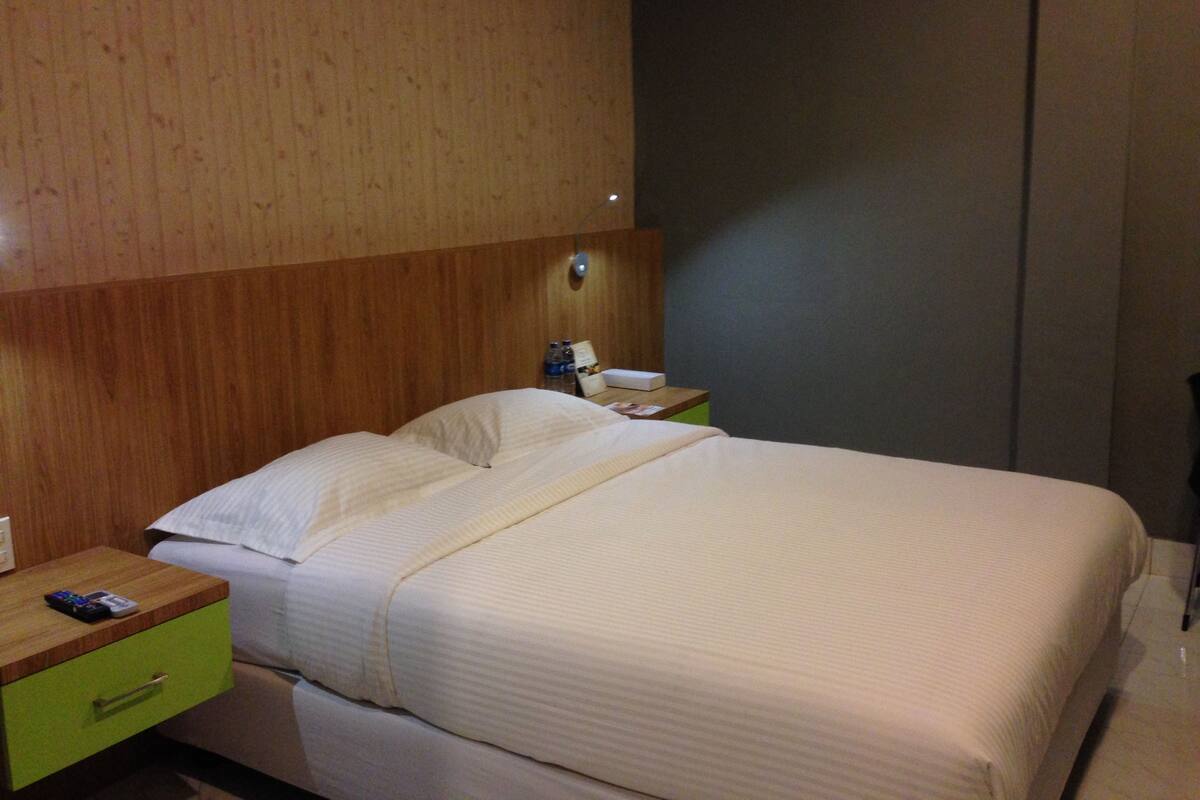 A hotel room with a bed, a bedside table with a remote control, a nightstand with a lamp, and a green drawer.