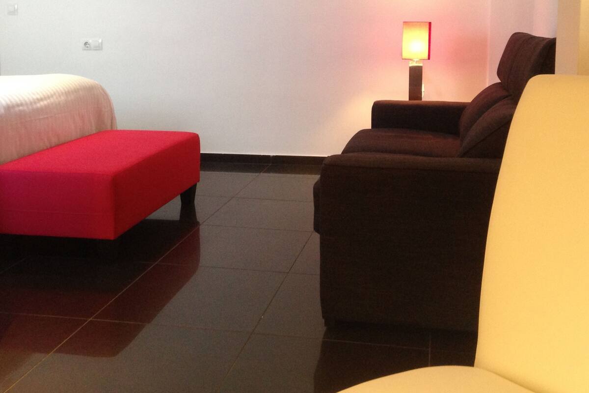 Una habitación con una cama, un reposapiés rojo, un sofá marrón y un sillón amarillo.