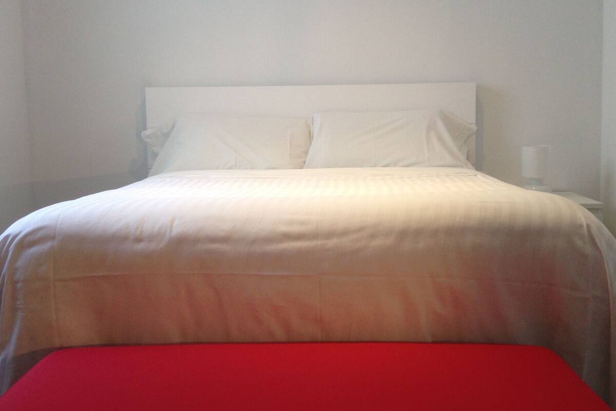 Una cama con ropa blanca y un banco rojo adelante.