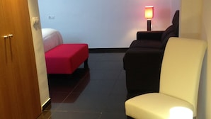 Habitación de hotel con cama, un pouf rojo, un sofá negro, un sillón blanco y una mesita de noche con lámpara.