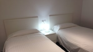 Una habitación con dos camas individuales, una mesita de noche con lámpara y pared blanca.
