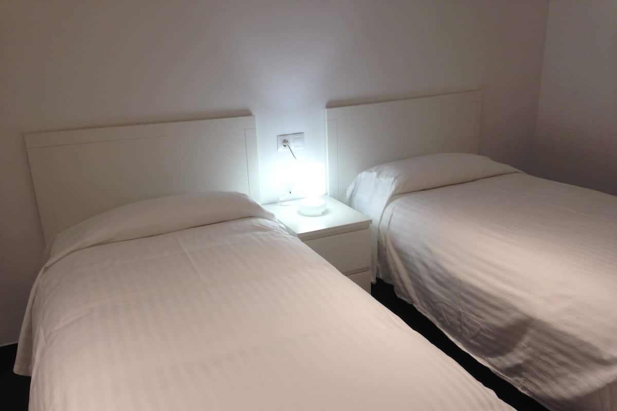 Una habitación con dos camas individuales, una mesita de noche con lámpara y pared blanca.
