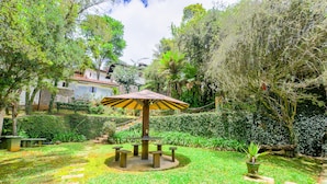 Un jardín con una mesa y sillas centrales, rodeado de abundante vegetación y una casa al fondo.
