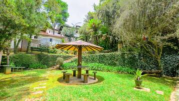Um jardim com uma mesa e cadeiras no centro, rodeado por vegetação exuberante e uma casa ao fundo.