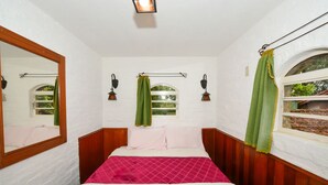 Un dormitorio con cabecera de madera, un espejo grande, dos lámparas de pared, una ventana con vistas, y una cama con edredón rosa y blanco.