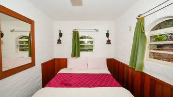 Quarto com cabeceira de madeira, um espelho grande, duas luminárias fixadas na parede, uma janela com vista e uma cama com colcha rosa e branca.