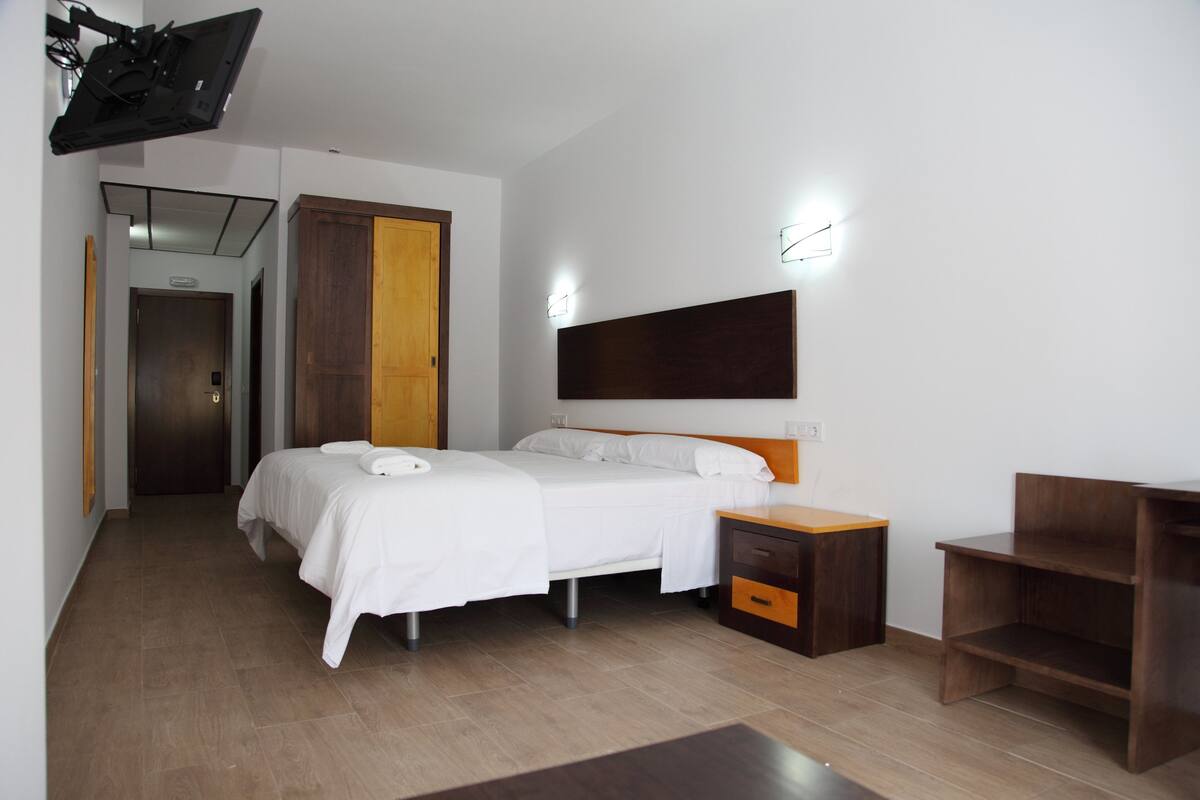 A hotel room with two beds, a wooden headboard, a nightstand, a desk, and a wardrobe.
