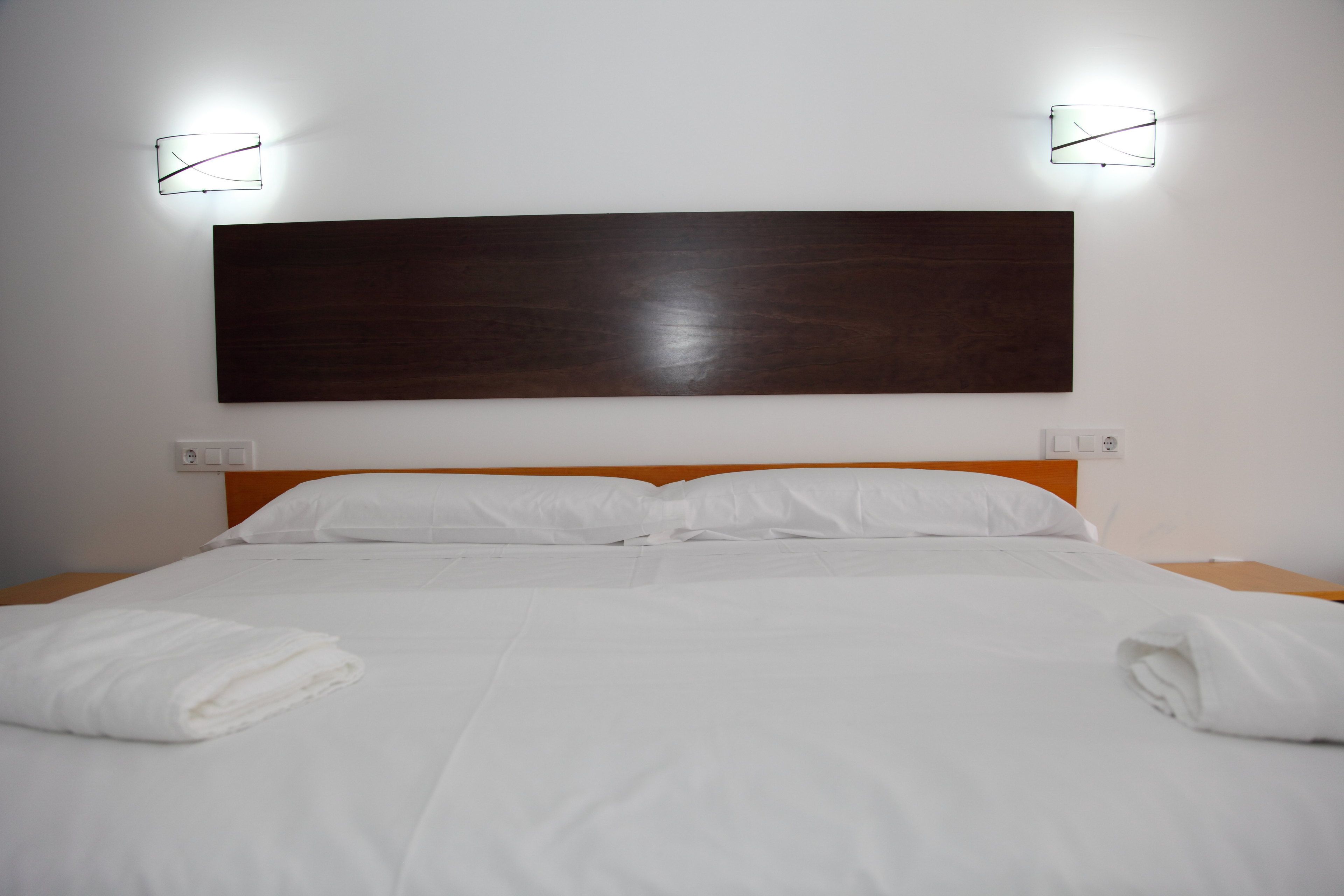A neatly made bed with white linens, a wooden headboard, and two wall-mounted light fixtures.