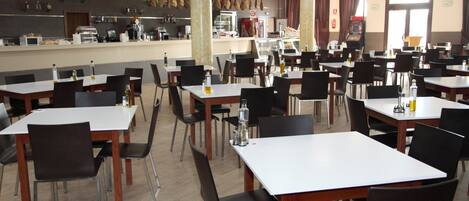 Un'ampia sala da pranzo con numerosi tavoli e sedie, un bancone con distributore di bevande e una zona cucina ben visibile.
