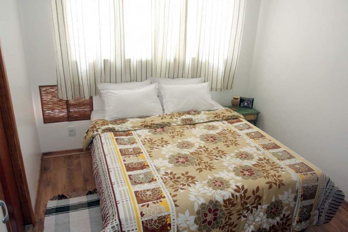 A bedroom with a bed, a nightstand, a window with curtains, and a wooden floor.
