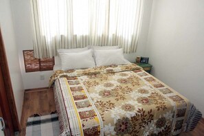 A bedroom with a bed, a nightstand, a window with curtains, and a wooden floor. - Pousada Vó Irene (Mangaratiba)