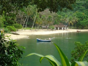 A serene lakeside scene with a small boat on the water, surrounded by lush greenery and a sandy beach with a small building. - Pousada Vó Irene (Mangaratiba)