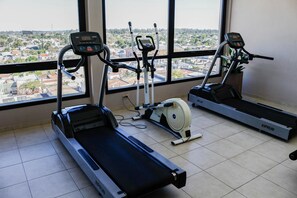 Ein Fitnessraum mit Laufbändern, einem Crosstrainer und einem Hometrainer, mit Blick auf eine Stadtlandschaft durch große Fenster.