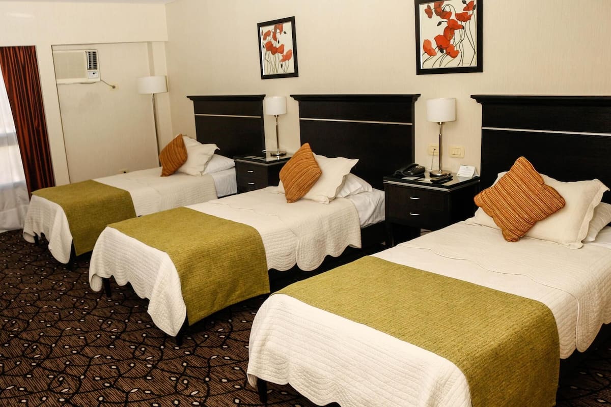 Une chambre d’hôtel avec trois lits simples, chacun recouvert d’une courtepointe blanche et d’un plaid vert. On y trouve deux tables de chevet avec des lampes, un climatiseur fixé au mur, et deux œuvres d’art florales encadrées au-dessus des lits.