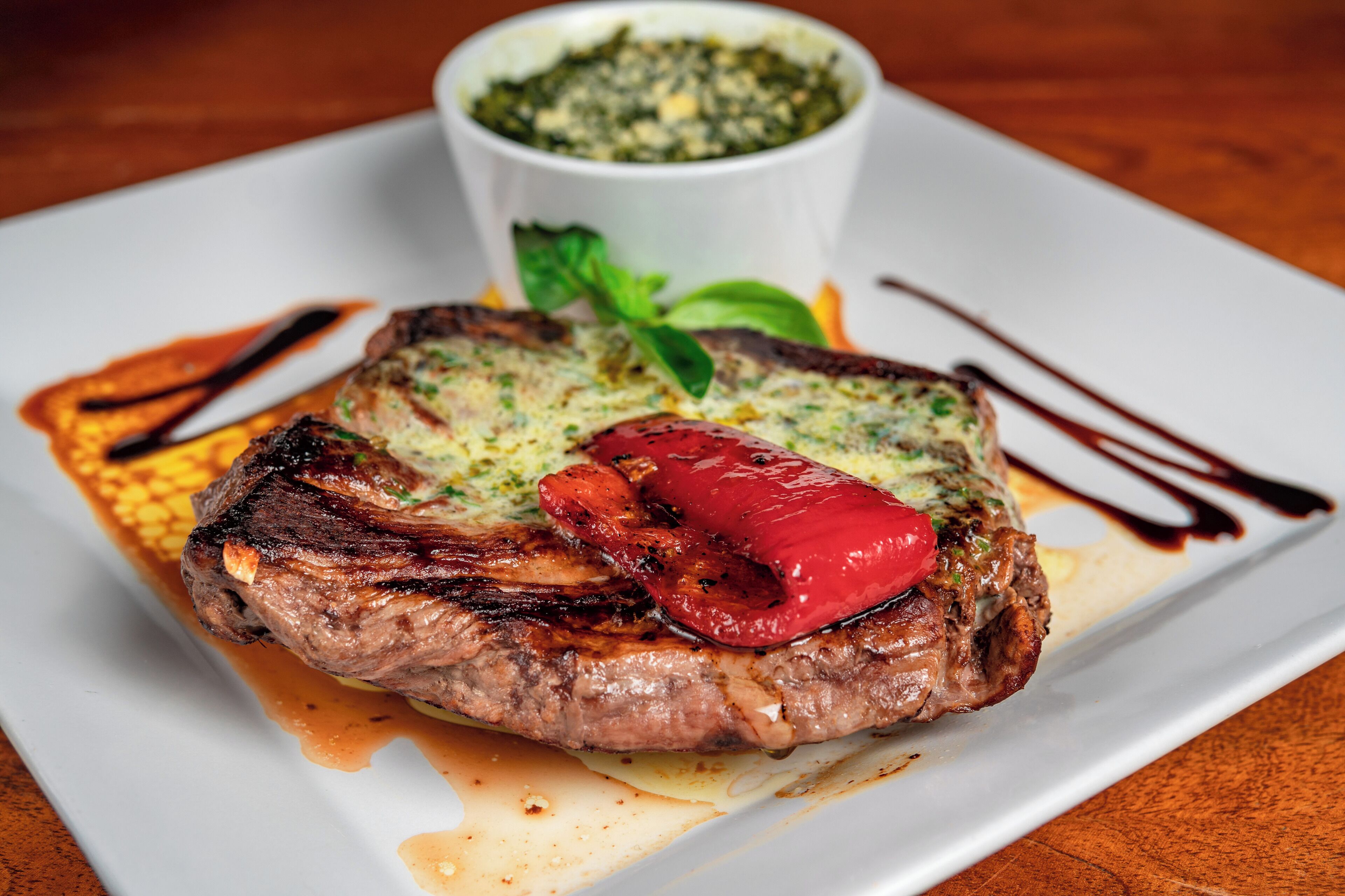 A steak with a grilled patty and a red pepper on a white plate, accompanied by a small bowl of sauce.