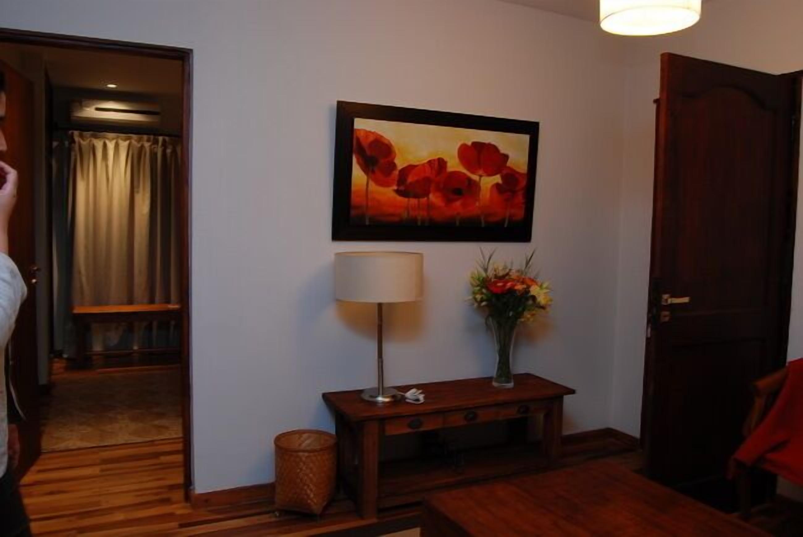 Una habitación con piso de madera, un cuadro enmarcado de flores rojas, una lámpara y un florero con flores sobre una mesita.